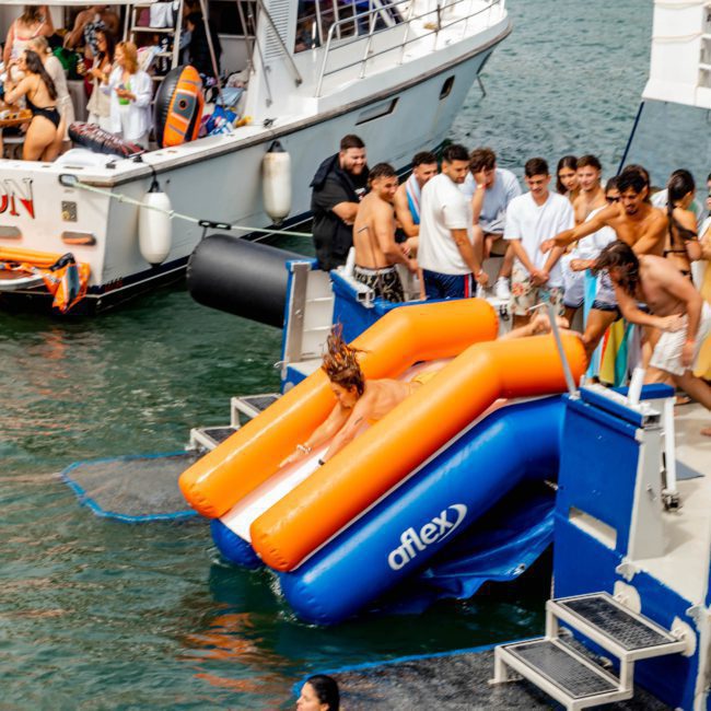 A group of people enjoying a Catamaran party Sydney. One person is sliding down an inflatable orange and blue slide into the water, while others eagerly wait their turn with the boats anchored in the beautiful Sydney Harbour.