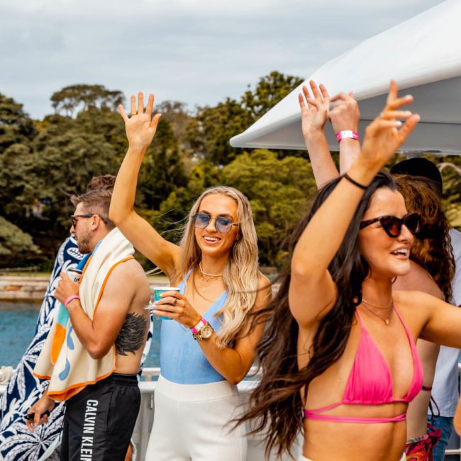 People are dancing and enjoying a corporate boat event under cloudy skies, with trees and water in the background. They are wearing casual summer attire, and some have towels draped over their shoulders.