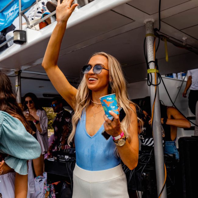 A woman wearing sunglasses is raising her hand and holding a drink at a lively outdoor event on a luxury yacht hire in Sydney. Other people are gathered around her, some on a boat deck, enjoying the sunny and bright weather.