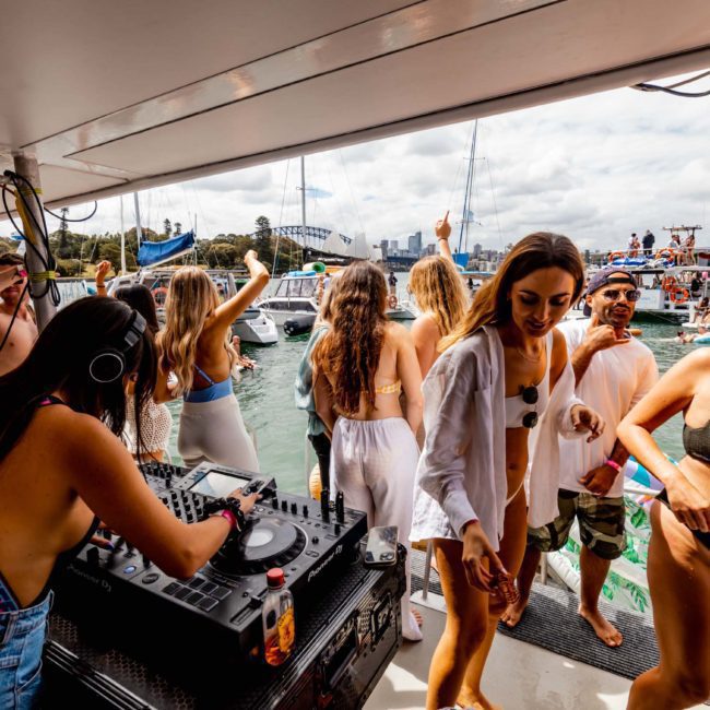 People are dancing and enjoying a sunny day on a luxury yacht hire in Sydney, with a DJ playing music. The background shows other boats on the water.