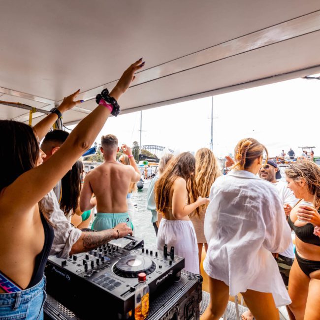 A group of people in swimwear dancing and socializing on a catamaran party Sydney, with DJ equipment visible in the foreground and other boats in the background.