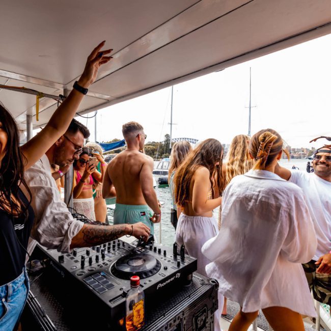A group of people dancing and enjoying a Sydney boat party hire on a luxury yacht, with a DJ playing music. The scene includes both men and women wearing casual and swim attire.