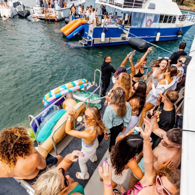 A group of people partying on a private yacht charter in Sydney Harbour with more boats and people in the background. Some are dancing, others are chatting, and inflatable toys float nearby, making it a perfect catamaran party in Sydney.