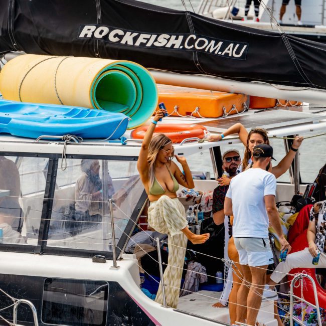 A group of people socializing on a boat with water sports gear. The boat has "ROCKFISH.COM.AU" written on it, and a woman is stepping onto the deck. Sydney's beautiful harbor sets the scene, perfect for luxury yacht hire or an unforgettable DJ boat party. Other boats are visible in the background.