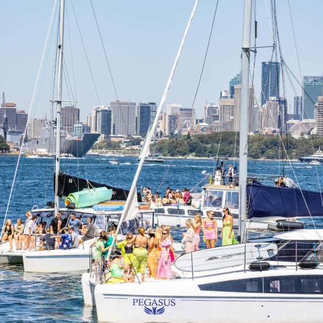 Several boats with people onboard are anchored on a body of water, with a cityscape in the background. One boat in the foreground, named "PEGASUS," stands out amidst luxury yacht hire Sydney options.