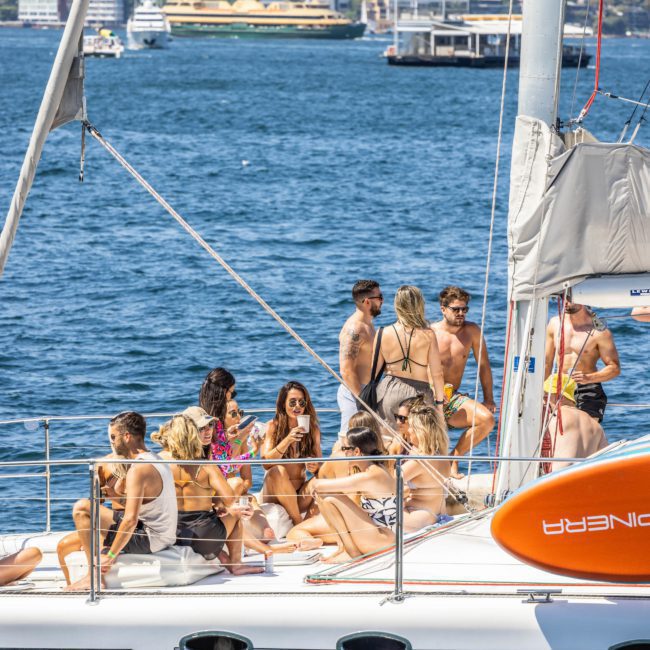A group of people socialize on the deck of a private yacht charter Sydney Harbour with buildings and other boats visible in the background.
