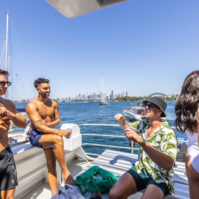 A group of people socialize on a luxury yacht hire in Sydney under clear blue skies with a city skyline in the background. One person is opening a bottle while others relax and chat.