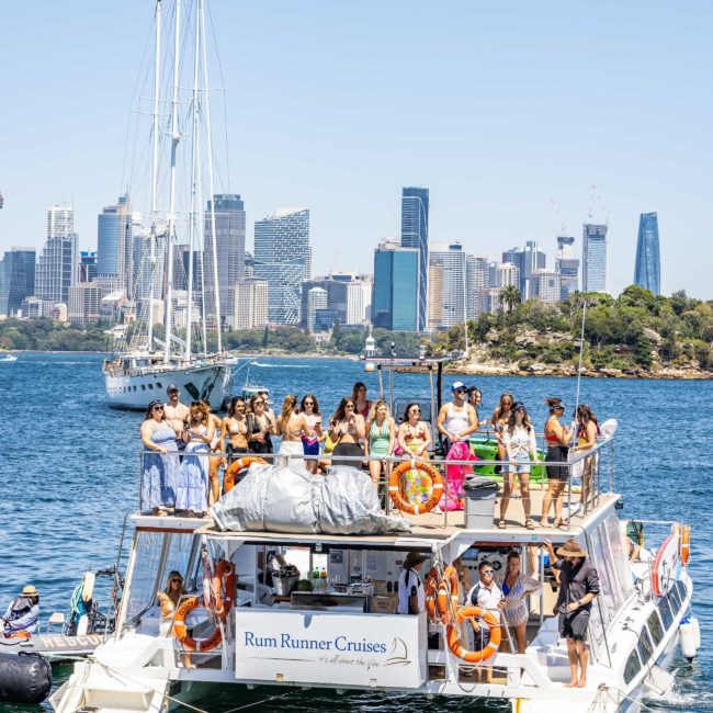A boat named "Rum Runner Cruises" with many people on board is sailing in a harbor with a cityscape and sailboats in the background, perfect for a Sydney boat party hire.
