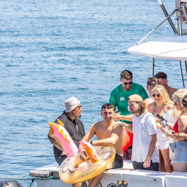 A group of people on a boat enjoying a sunny day. One man has a colorful pool float shaped like an octopus. The boat, named "WACKATO," is part of a Sydney boat party hire with sparkling blue water in the background.