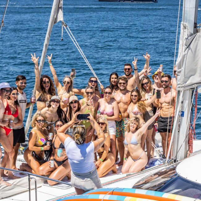 A group of people in swimwear pose for a photo on a private yacht charter in Sydney Harbour under sunny skies.