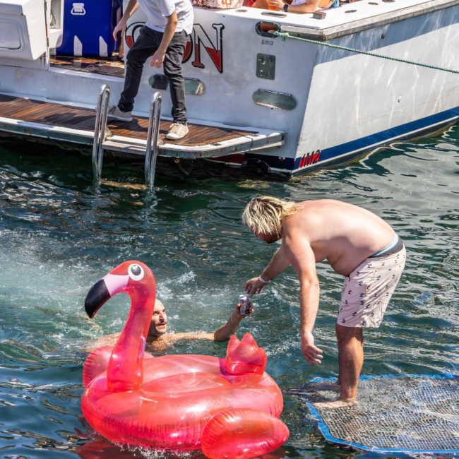 A person on a private yacht charter in Sydney Harbour pours a drink into another's mouth, who lounges on a large inflatable pink flamingo in the water, surrounded by friends.