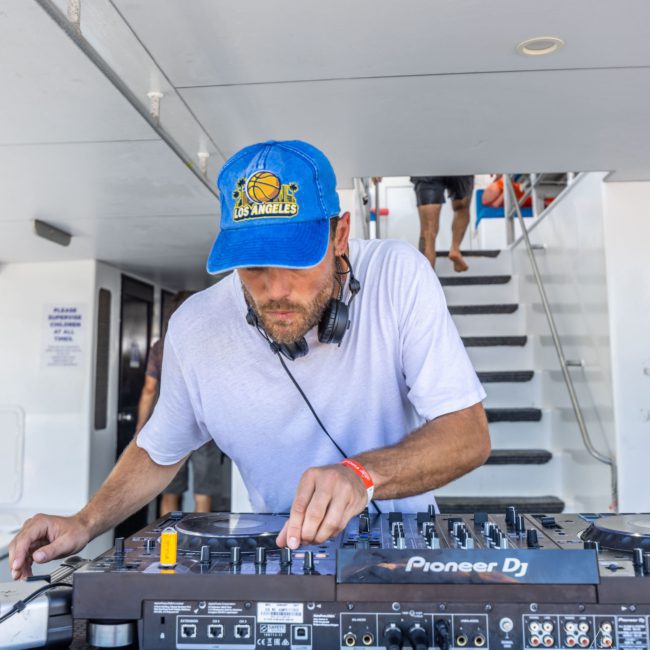 A DJ wearing a blue Los Angeles cap and white t-shirt is focused on operating a Pioneer DJ mixer with headphones around his neck. There are stairs and a few people in the background, adding to the lively atmosphere of a Sydney boat party hire.