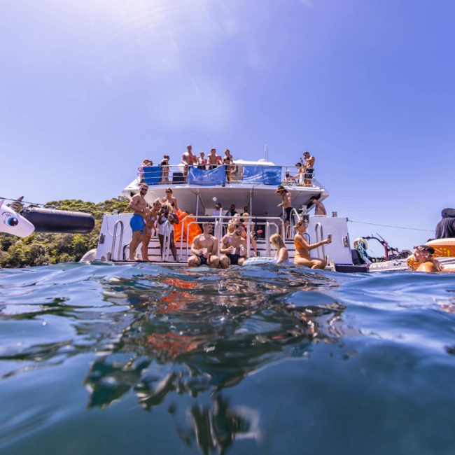 A group of people are swimming and floating on inflatables in the water near a docked boat, with others gathered on the boat under a clear blue sky, enjoying a lively catamaran party Sydney.
