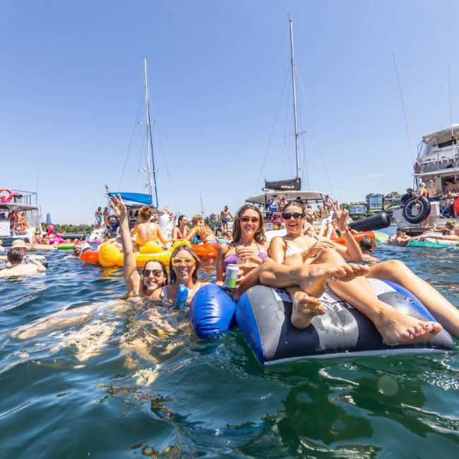 A group of people relax on inflatable loungers in the water, surrounded by boats, enjoying a sunny day. It’s the perfect setting for a Sydney boat party hire or catamaran party Sydney.