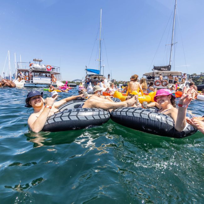People relaxing on inflatable tubes in a crowded body of water on a sunny day, surrounded by boats and other swimmers, with some individuals holding drinks, while a DJ boat hire Sydney keeps the vibe lively.