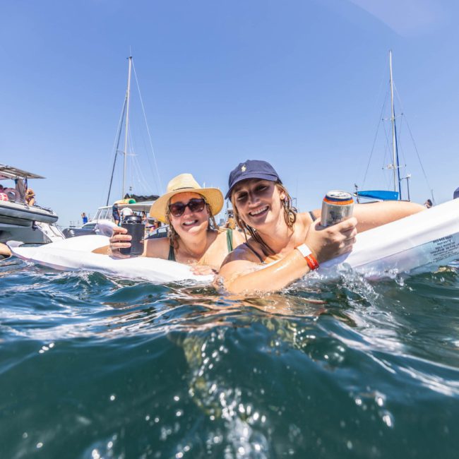 Two people enjoy drinks while floating on a raft in the water near boats during a sunny day, embracing the vibe of a luxury yacht hire in Sydney.