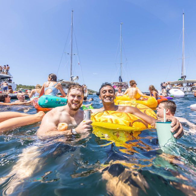 People enjoy a sunny day in the water, holding drinks and inflatables, with boats and more people in the background. For an unforgettable experience, consider a catamaran party in Sydney or luxury yacht hire.