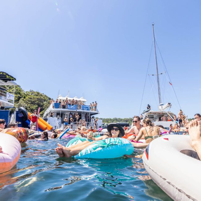 People are lounging on inflatable rafts in a body of water surrounded by boats on a sunny day, enjoying the atmosphere of a corporate boat event in Sydney.