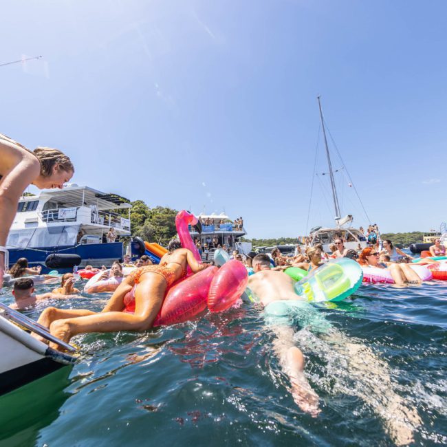 People enjoying a Catamaran party Sydney on the water with boats and inflatables under a clear, sunny sky.