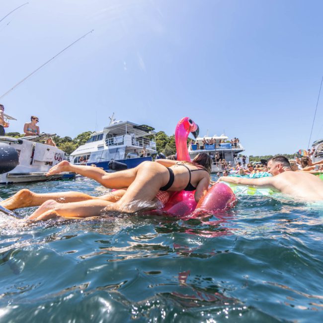 People enjoying a sunny day on boats and in the water, some floating on inflatable toys, at a crowded marina. The scene is lively with groups celebrating aboard private yacht charters and catamaran parties around Sydney Harbour.