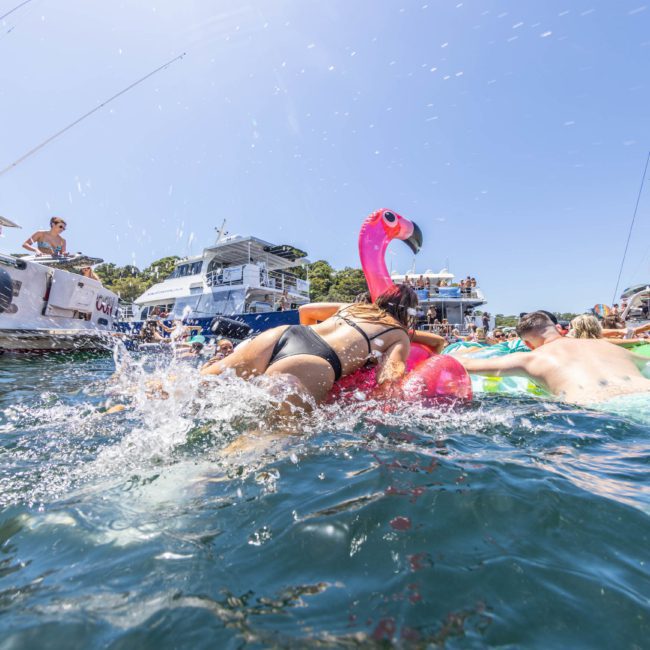 People swimming and lounging on inflatable floats in a crowded water area surrounded by boats on a sunny day, enjoying the atmosphere of a private yacht charter in Sydney Harbour.