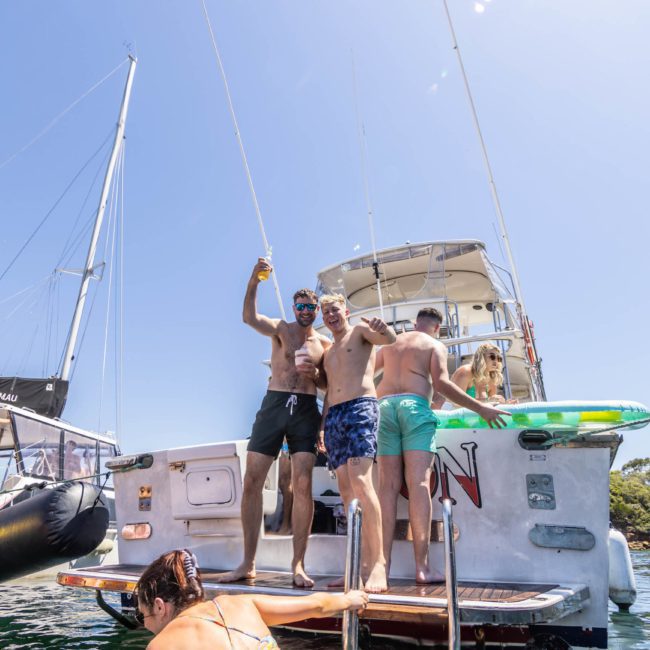 A group of people stands on the back of a boat, holding drinks and socializing, with another boat anchored nearby. Some individuals in swimwear are enjoying the sunny day on the water, making it a perfect scene for a Sydney boat party hire.