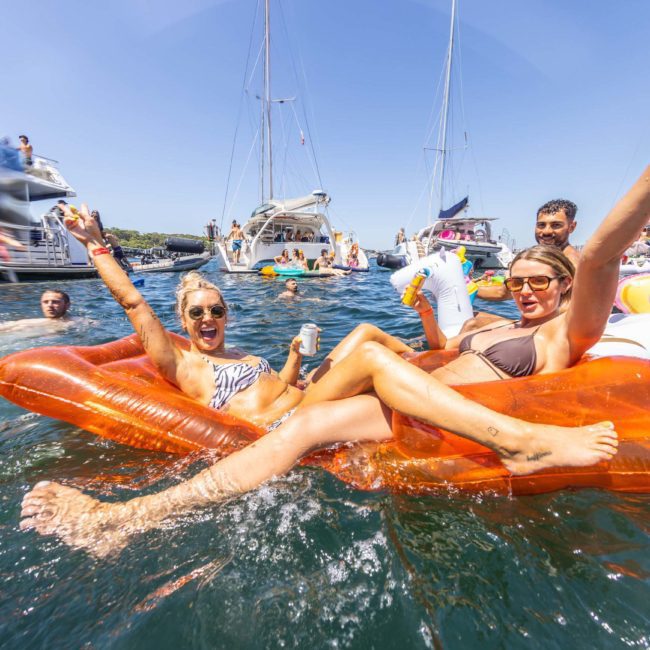 People on orange inflatable lounge chairs enjoying a catamaran party in the water surrounded by boats and other floatation devices. The event is characterized by a lively atmosphere under clear skies.