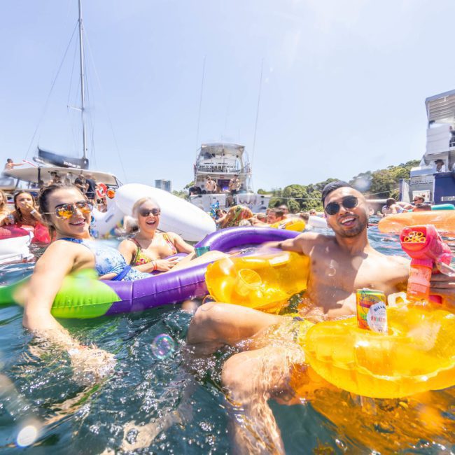 People are lounging on inflatable floats in the water, enjoying a sunny day. Various boats, including a private yacht charter Sydney Harbour, are in the background.