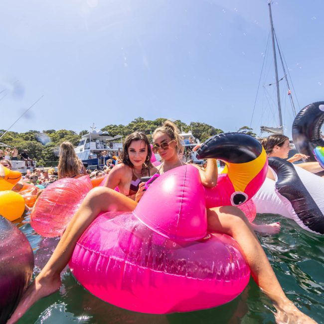 A group of people enjoying a sunny day on inflatable pool floats in the water, with boats and greenery visible in the background, basking in the fun atmosphere of a Sydney boat party hire.