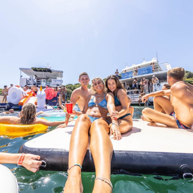 A group of young people in swimsuits are relaxing on a floating platform in the water near two boats, with more people socializing and swimming around them on a sunny day. They enjoy the vibrant ambiance of a Catamaran party Sydney, making it an unforgettable experience.