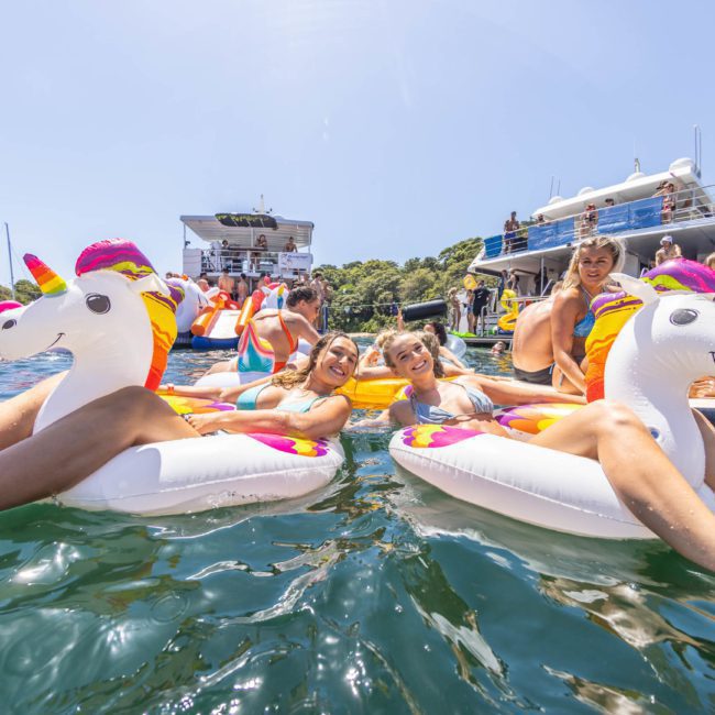 People enjoying a sunny day on the water, lounging on inflatable unicorn floats surrounded by boats and other flotation devices. In the distance, a luxury yacht hire Sydney offers an elegant backdrop for their fun.