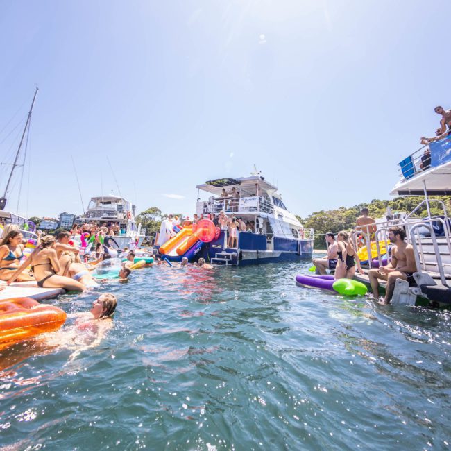 People are enjoying a sunny day on a lake, swimming and lounging on inflatable rafts near boats that are docked together. It’s a perfect setting for those considering luxury yacht hire in Sydney or perhaps even a vibrant catamaran party in Sydney.