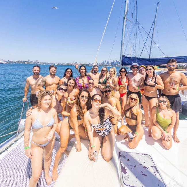 A group of people in swimwear are gathered on a luxury yacht on a sunny day, with the stunning Sydney city skyline visible in the background.