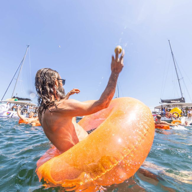 A person with long hair, wearing sunglasses and an orange inflatable ring, tosses a ball while in the water with boats and other people on inflatable rings in the background, enjoying a private yacht charter on Sydney Harbour.