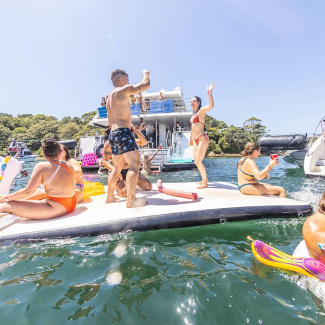 A group of people in swimsuits enjoy a sunny day on a large floating platform on the water, surrounded by boats. Some are sitting, others standing and dancing, while various inflatables float around. It's the epitome of a Catamaran party Sydney.