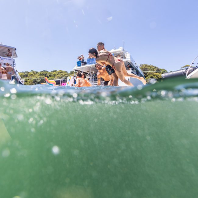 A group of people having a fun time swimming and jumping from a yacht on a sunny day, with the lower half of the image showing the water and the upper half showing part of the yacht and people enjoying a lively catamaran party in Sydney.