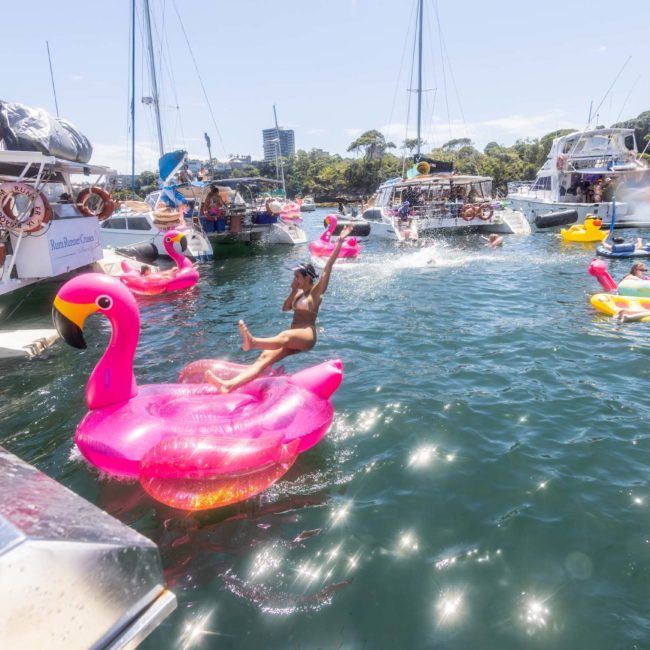 People enjoying a sunny day on the water with inflatable toys and boats. A person is jumping towards a pink flamingo float. Several boats and other inflatables, including luxury yacht hire Sydney options, are visible in the background.