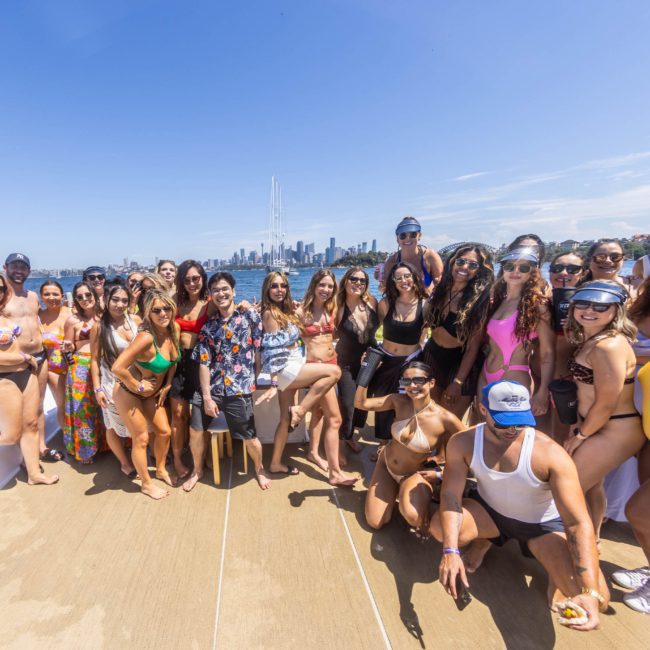 A large group of people in swimwear pose for a photo on a private yacht charter Sydney Harbour, with a city skyline visible in the background.