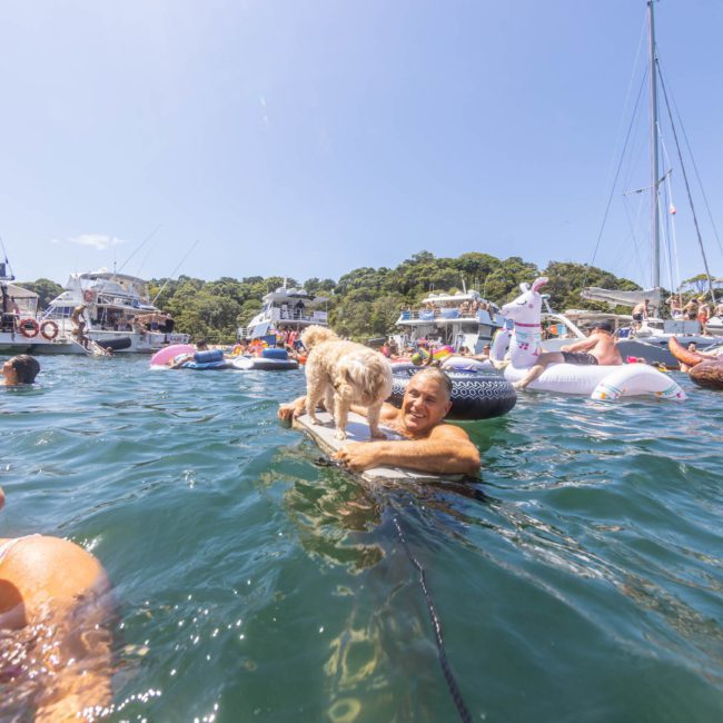 People enjoying a sunny day on the water, swimming and relaxing on boats and floaties, with a dog standing on a floating raft. Amidst this idyllic scene, a private yacht charter in Sydney Harbour adds an extra touch of luxury.