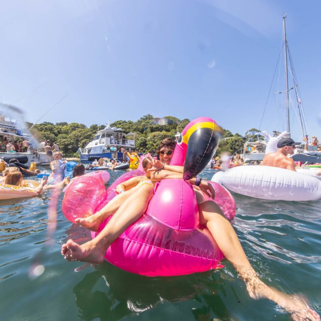 People are enjoying a sunny day on various inflatable floats in a busy water area, surrounded by boats. A person is prominently seated on a large pink flamingo float, while nearby, others revel in the music from a DJ boat hire Sydney.