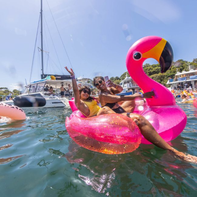 Two people are floating on an inflatable pink flamingo in a scenic water area with boats and other inflatables nearby. Both are waving and appear to be enjoying the moment, while a stunning private yacht charter cruises by in Sydney Harbour.