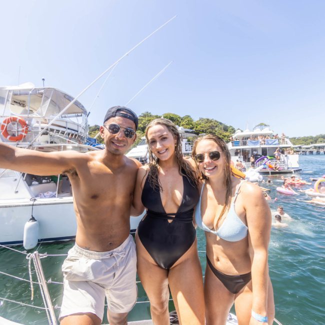 Three people in swimwear stand on a luxurious yacht, smiling at the camera with other boats and inflatables visible in the background on a sunny day.