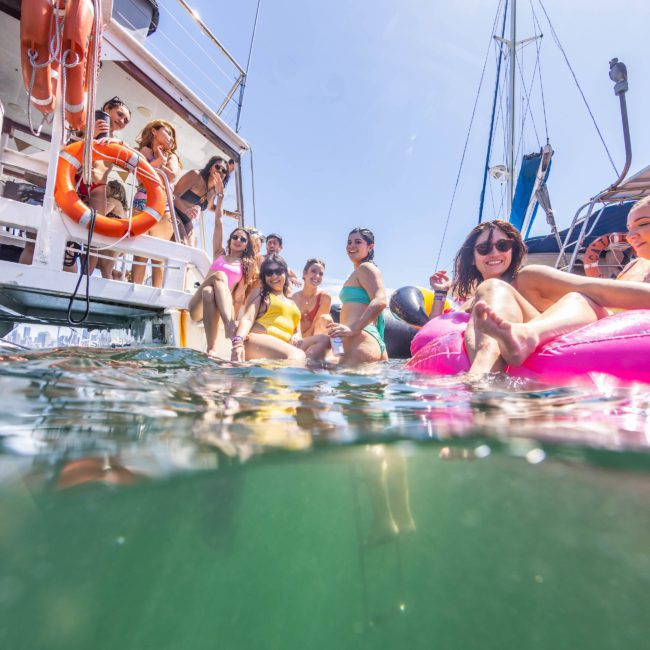 A group of people in swimsuits are enjoying a vibrant Sydney boat party hire on the water. Some are on the luxurious yacht, some are swimming, and one person floats on a pink inflatable.