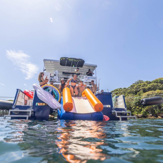 A group of people enjoy a sunny day on a large boat with an attached inflatable slide, floating in clear water near a forested area. Some are mingling on the deck while others have fun on the slide, making it an ideal scene for luxury yacht hire Sydney.