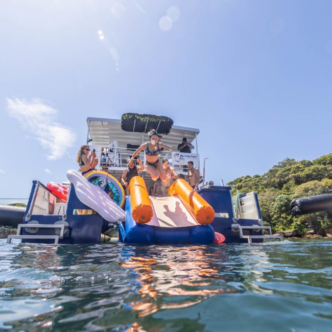 People enjoying a sunny day on a catamaran party in Sydney, with one person sliding into the water and others standing or sitting nearby. The surroundings include greenery and other boats, perfect for a memorable Sydney boat party hire.