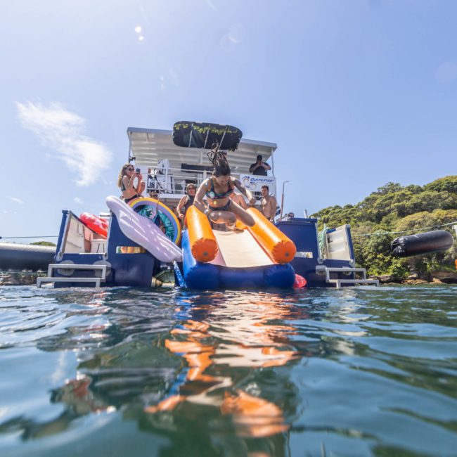 Person slides down an inflatable slide attached to a luxury yacht hire Sydney, with other people on the boat and in the water around. Bright, sunny day with clear water and green trees in the background.