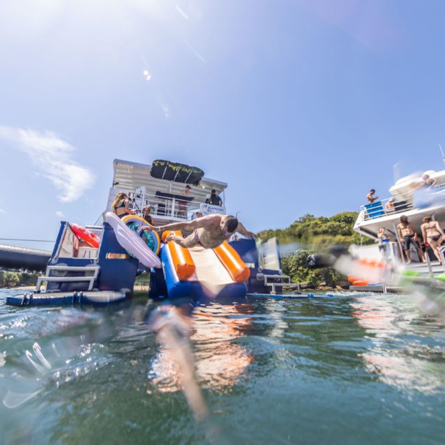 A person jumps off an inflatable slide into the water from a boat, with other people on nearby boats watching under a clear, sunny sky. This scene captures the essence of private yacht charters in Sydney Harbour.