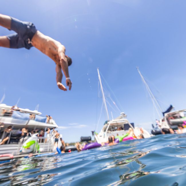 A person in swim trunks is diving into the water from a boat, with several boats and people in the background under a clear blue sky, enjoying the lively atmosphere of a Sydney boat party hire.