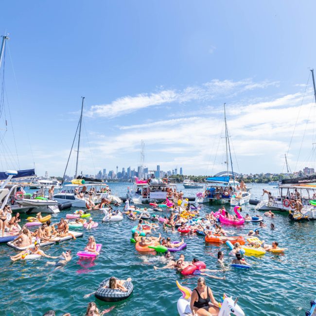 A large group of people relax on colorful inflatable floats and swim near boats in a sunny harbor with a cityscape in the background, enjoying a memorable DJ boat hire Sydney experience.