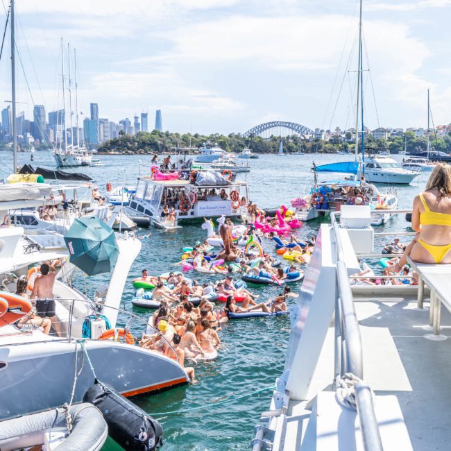 Numerous people on boats and inflatables gather in a bustling waterway near a city skyline and iconic bridge on a sunny day, making it perfect for a Sydney boat party hire or even considering a private yacht charter Sydney Harbour.